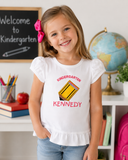 Young girl wearing a personalized white ruffle shirt with embroidered Kindergarten, Light Gold pencil appliqué, and the name Kennedy in a classroom setting