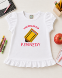 Flat lay of personalized white girls’ ruffle shirt with Kindergarten embroidery, Light Gold pencil appliqué, and name Kennedy