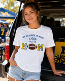 Woman at a football tailgate wearing a white personalized Football Mom shirt with AYDEN, blue-and-gold M letters, and a real leather football appliqué.