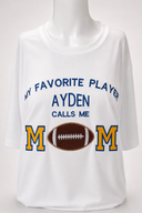 White personalized Football Mom shirt on a female mannequin with AYDEN, blue embroidery, gold-and-blue M letters, and a real brown leather football.
