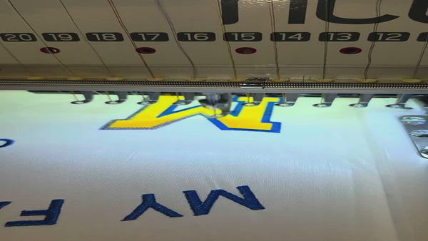 Embroidery machine stitching blue-and-gold tackle-twill M letters onto a white personalized Football Mom shirt.